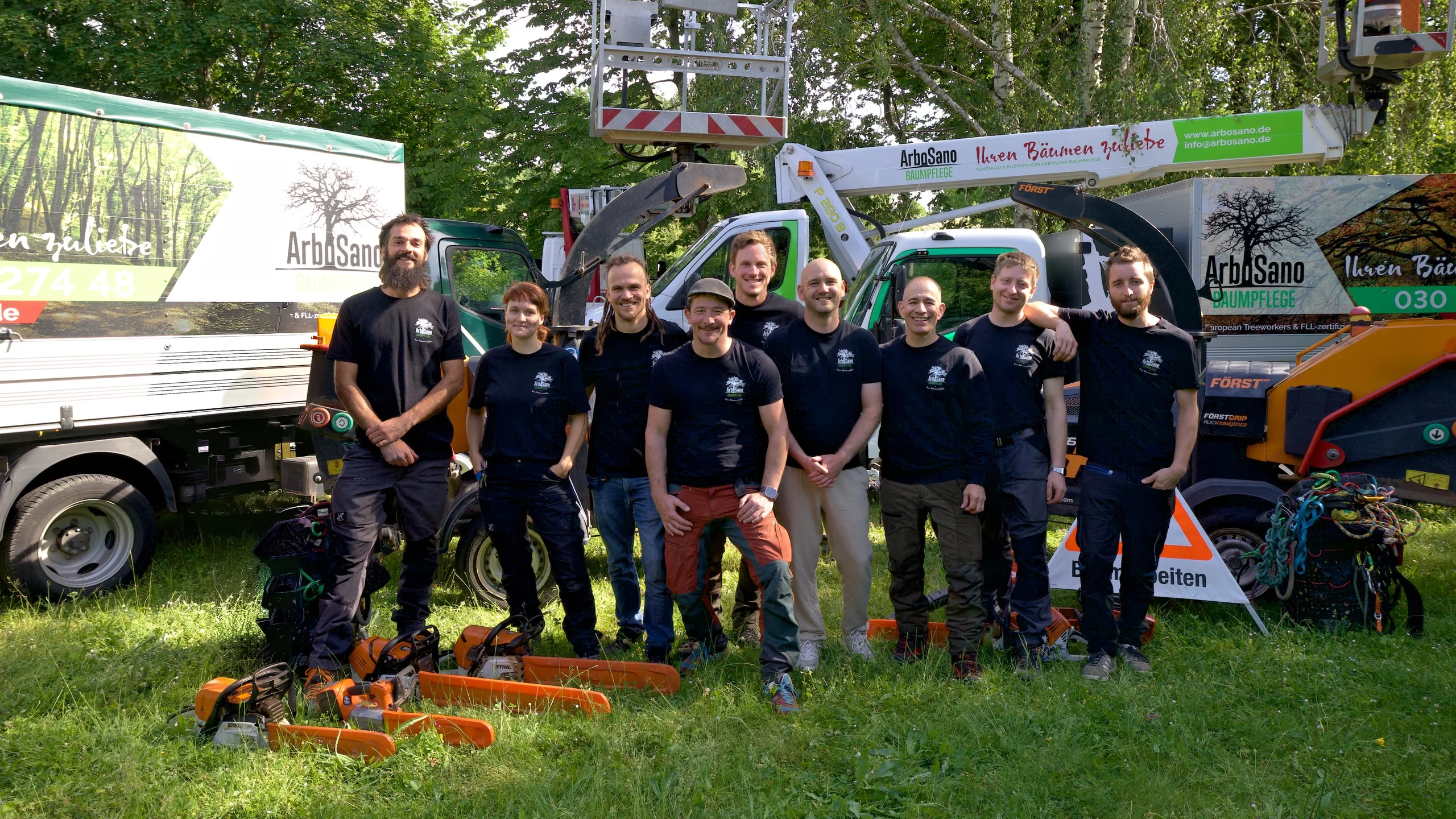 Das Team von ArboSano in Arbeitskleidung vor dem Fuhrpark mit Hubarbeitsbühne, Häcksler und Motorsägen
